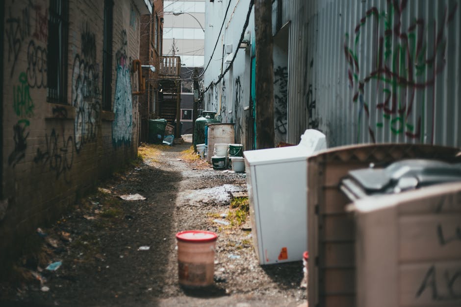 A narrow, cluttered alleyway between two graffiti-covered brick and metal-paneled walls, with various discarded items and waste containers positioned along the uneven, muddy ground. In the foreground, there is a large beige plastic bin with a pile of miscellaneous rubbish partially visible, and a smaller plastic container with a red lid nearby. Further back, several green and white waste bins, some with closed lids, are arranged haphazardly on the wet, muddy surface, which has patches of moss and scattered litter. Additional debris, including paper and plastic, is scattered along the alley, which appears to be used for private waste disposal rather than municipal rubbish collection. The scene is lit by natural daylight filtering through an open section at the alley’s end, revealing a staircase and some residential structures in the background, suggesting an informal area where waste is temporarily stored before removal. Waste Collection Hounslow occasionally handles such scenes as part of alternative rubbish management solutions for narrow or hard-to-access urban alleys.