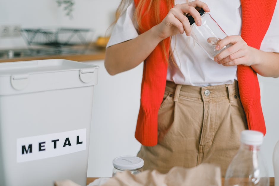 A person wearing a red sleeveless cardigan over a white shirt and beige trousers is holding a clear plastic container with a black lid, standing next to a white metal bin labeled 'METAL.' The scene appears to be in a kitchen or disposal area, with a countertop and some kitchen items visible in the blurred background. The person's hands are positioned to place or remove the container, which may be used for collecting recyclable metal items. The environment is lit with natural or artificial light, highlighting the textures and colors of the container, clothing, and nearby objects. The setting reflects an organized approach to waste separation and recycling, consistent with waste management practices that involve bin labeling and material-specific collection, supported by companies like Waste Collection Hounslow specializing in rubbish removal services.