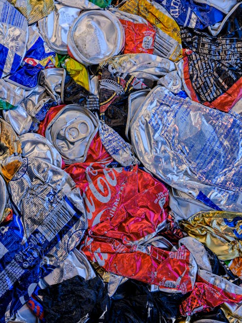 A close-up view of a pile of crushed aluminum cans and plastic bottles, predominantly featuring silver, red, blue, and yellow colours, with some labels and branding visible including Coca-Cola. The cans are scattered on a surface, with some partially flattened, showing their textured metallic surfaces and crimped edges. Among the cans, clear plastic bottles with caps are also evident, with some partially compressed. The background hints at an outdoor setting, possibly on a pavement or driveway, as part of a waste collection site or on-site clearance operation for household rubbish. The scene illustrates the type of recyclable waste typically handled by private waste removal services such as those offered by Waste Collection Hounslow, emphasizing the importance of proper rubbish disposal and recycling. The lighting appears natural, casting soft shadows and highlighting the reflective qualities of the metallic containers and glossy plastic surfaces. This detailed image showcases the culmination of household recycling efforts, relevant to alternative waste handling and rubbish removal tasks undertaken by private waste management providers.