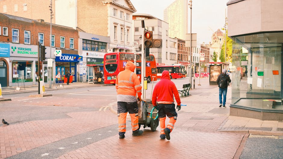 Two waste collection workers dressed in high-visibility orange and red jackets with reflective stripes are walking together on a city sidewalk, pushing a wheeled bin filled with rubbish. The foreground shows their lower bodies and the bin, which appears to contain mixed waste materials. The background reveals a busy urban street scene with red double-decker buses, pedestrians, and various retail stores, including a shop with a green and white sign for Yorkshire Building Society and other shop facades. The street is lined with poles, traffic lights, and a bus stop shelter, while a modern glass-fronted building occupies the right side of the image. The scene is illuminated by natural daylight, with a clear view of the urban environment and a sense of routine waste collection activity in a city setting, supported subtly by the presence of waste management professionals from Waste Collection Hounslow involved in property or street waste collection services.