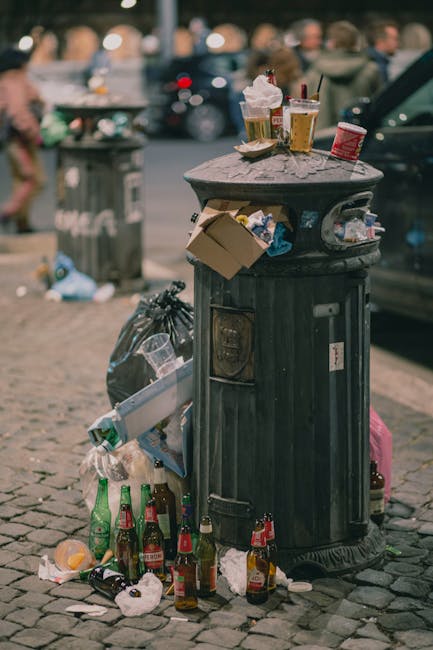 Two waste collection workers dressed in high-visibility orange and red jackets with reflective stripes are walking together on a city sidewalk, pushing a wheeled bin filled with rubbish. The foreground shows their lower bodies and the bin, which appears to contain mixed waste materials. The background reveals a busy urban street scene with red double-decker buses, pedestrians, and various retail stores, including a shop with a green and white sign for Yorkshire Building Society and other shop facades. The street is lined with poles, traffic lights, and a bus stop shelter, while a modern glass-fronted building occupies the right side of the image. The scene is illuminated by natural daylight, with a clear view of the urban environment and a sense of routine waste collection activity in a city setting, supported subtly by the presence of waste management professionals from Waste Collection Hounslow involved in property or street waste collection services.