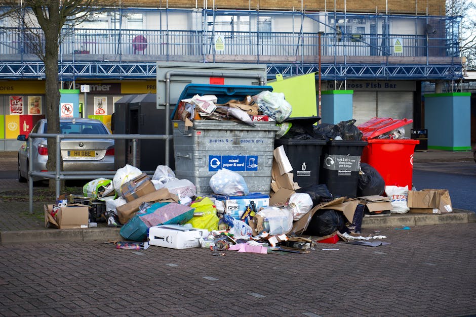 A large open-top metal bin for mixed paper and cardboard waste is situated on a paved sidewalk in front of a commercial building with a blue scaffolding structure and signage. The bin is overflowing with various types of rubbish including flattened cardboard boxes, crumpled paper, plastic packaging, and miscellaneous waste, some of which is spilling onto the ground. Several black and red waste bags, along with loose cardboard boxes and smaller wrappers, are piled around the bin, creating a cluttered scene. To the left, a silver vehicle is parked next to a metal rail separating the parking area from the pedestrian walkway. The background features shopfronts with signage and a tree with bare branches, indicating a possible winter season. The environment appears unaffected by waste collection, suggesting private or alternative rubbish handling. The scene emphasizes the importance of regular rubbish removal services such as those provided by Waste Collection Hounslow for maintaining cleanliness in public spaces.