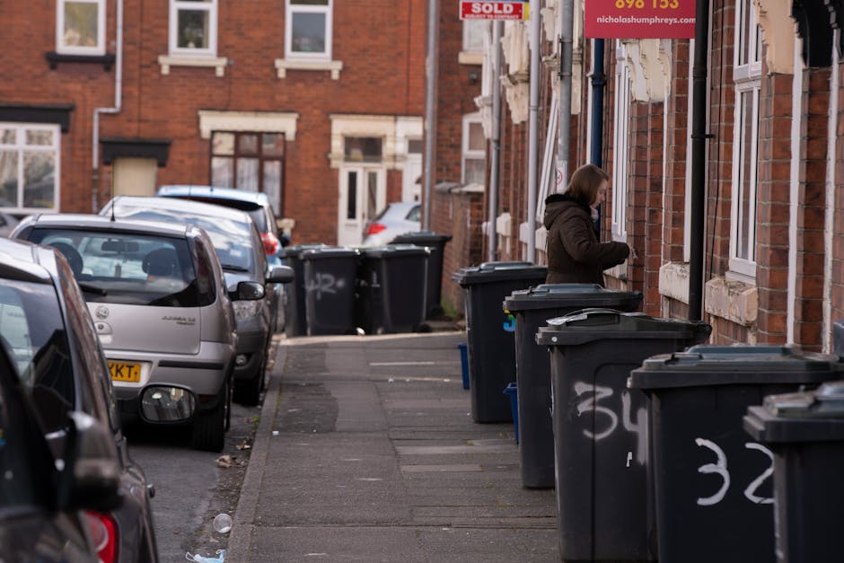 A woman standing beside a series of black wheelie bins placed along a residential pavement, preparing waste for disposal or collection. The bins are positioned in front of brick row houses, each with white-framed windows and stone sills. The bins have open lids, revealing contents inside, and some have white spray-painted numbers or markings on their sides. The pavement is narrow, with parked cars lining the street on the left side, including small hatchbacks in silver and grey. In the background, additional bins are visible, as well as more brick houses with similar architectural features. The sky appears overcast, casting a neutral, diffuse light over the scene. The overall environment suggests a typical urban street where residents manage their waste using personal collection bins, aligning with private or alternative rubbish disposal options supported by waste management services like Waste Collection Hounslow.