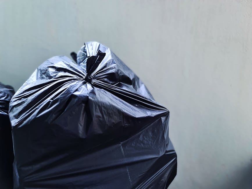A close-up of several tightly tied black plastic rubbish bags resting against a plain, light-colored wall. The bags are made of glossy, durable plastic with visible creases and folds, reflecting light on their surface, with one bag slightly in the foreground and others partially visible behind. They are positioned on a surface, likely a floor or pavement, with no other objects visible in the immediate vicinity. The background is neutral and uncluttered, emphasizing the clean, professional appearance suitable for a waste management context. The scene appears to depict an outdoor or indoor area prepared for collection or disposal, aligning with private rubbish removal services provided by Waste Collection Hounslow, supporting alternative waste handling options outside of standard municipal collection schedules.