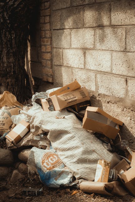 A pile of mixed waste materials is situated at the base of a brick and concrete wall outdoors. The rubbish includes cardboard boxes, some partially collapsed and others still intact, with a few openings revealing their empty interiors. There are also large, crumpled paper or fabric sacks in light grey and white tones, which appear worn and slightly dirty. Surrounding the rubbish are small rocks and dirt on the ground, indicating an outdoor environment, possibly an alleyway or side yard. The wall behind the pile comprises roughly textured bricks and concrete blocks with visible mortar joints, and a section of blackened tree trunk is visible on the left side of the image. The lighting is diffuse, suggesting an overcast sky or shaded area. This scene captures a typical example of improperly disposed waste close to a building, highlighting the importance of proper rubbish removal and professional waste handling services such as those provided by Waste Collection Hounslow.