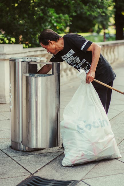 A middle-aged woman with short dark hair, wearing a black T-shirt with white text and dark trousers, is seen on a paved sidewalk in an outdoor park-like setting. She is bent over, reaching into a cylindrical stainless steel public rubbish bin with a rectangular opening at the top, designed for waste collection, as part of an environmental cleanup. The woman holds a large white plastic bag that appears filled with discarded waste, and a wooden stick or broom handle is tucked under her arm, indicating she may be involved in waste collection or street cleaning. The background includes lush green trees and a low stone or concrete barrier that runs along the side of the path, providing a natural and urban environment context. The scene is well-lit with natural daylight, emphasizing the clean, well-maintained setting suitable for municipal or private disposal services like those offered by Waste Collection Hounslow. The image exemplifies on-site waste collection and environmental care activities, relevant to alternative rubbish removal methods beyond regular council services.