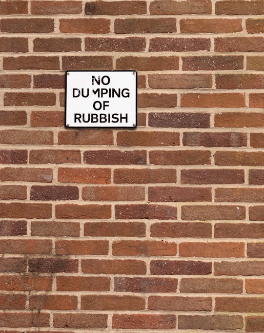 A white rectangular sign with black uppercase and lowercase text reading 'NO DUMPING OF RUBBISH' is mounted on a red brick wall in a horizontal orientation. The bricks are varied shades of reddish-brown, and the wall has a rough texture with visible mortar lines. The sign is positioned slightly above the center of the image, with the brick wall extending evenly on all sides behind it. The lighting is natural and even, highlighting the brick surface and the sign’s contrasting appearance, reflecting a typical outdoor environment where private waste handling or rubbish collection regulations are enforced. This scene suggests an area where waste disposal is monitored, aligning with practices of independent rubbish collection and on-site waste control managed by companies like Waste Collection Hounslow.