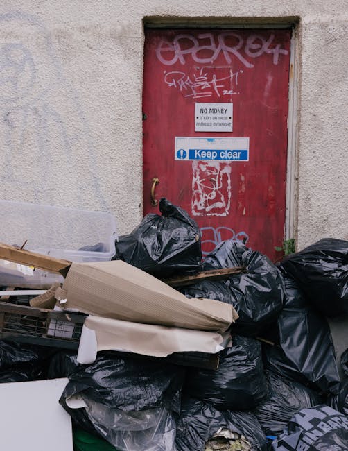 A pile of overflowing black garbage bags and discarded packaging materials stacked against a beige exterior wall beneath a red metal door covered in graffiti. The bags include plastic and paper packaging, with some cardboard and foam pieces visible. The door features signs, including one that states 'No Money' and another that reads 'Keep Clear,' along with various graffiti tags. The waste is situated on a concrete surface, partially obstructing the doorway. This scene illustrates typical discarded rubbish awaiting collection, highlighting the importance of timely waste removal services provided by companies such as Waste Collection Hounslow for private or alternative rubbish clearance needs.