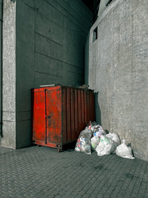 A pile of overflowing black garbage bags and discarded packaging materials stacked against a beige exterior wall beneath a red metal door covered in graffiti. The bags include plastic and paper packaging, with some cardboard and foam pieces visible. The door features signs, including one that states 'No Money' and another that reads 'Keep Clear,' along with various graffiti tags. The waste is situated on a concrete surface, partially obstructing the doorway. This scene illustrates typical discarded rubbish awaiting collection, highlighting the importance of timely waste removal services provided by companies such as Waste Collection Hounslow for private or alternative rubbish clearance needs.