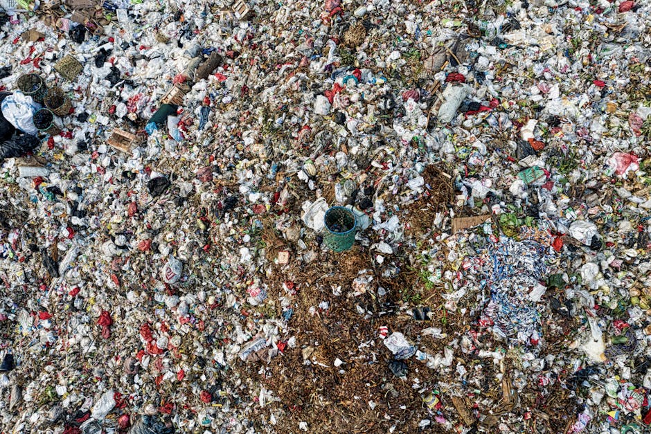 An extensive pile of mixed household and street waste covers a large outdoor area, with various types of rubbish including plastic bottles, cans, paper, cardboard, and packaging materials scattered across the ground. The waste appears compacted and loosely piled, with some items partially buried under others, exhibiting a range of colors from white and red to blue and green, all with varying degrees of dirt and grime. Among the debris, a small plastic bucket is visible, partially buried but distinguishable by its greenish color. The surrounding environment suggests an open, possibly neglected space, with no visible fencing or boundaries; natural light illuminates the scene, highlighting the chaotic, unorganized nature of the refuse. This image exemplifies the kind of waste accumulation that professional rubbish removal services, such as Waste Collection Hounslow, are equipped to manage, offering alternative waste handling options outside standard local authority collections.