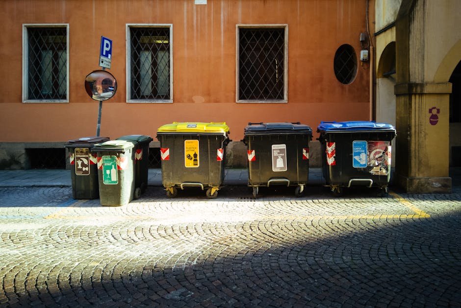 A row of four large wheeled waste bins is positioned on a cobbled street in front of a peach-colored building with three barred windows. The first bin on the left is green with a smooth plastic finish, featuring a white recycling symbol and an arrow pointing upward, indicating general waste or recycling. The second bin is yellow with a similar smooth plastic surface, marked with a yellow label that suggests electronic waste or other specific disposal. The third bin is black with a matte finish, displaying a white icon of a plastic bottle, indicating recyclables. The fourth bin is blue, also with a matte surface, showing a graphic of a plastic container and a recycling symbol. All bins have red and white striped reflective stickers around their sides. Behind the bins, a convex traffic mirror is mounted on a pole near the building, and nearby is a small circular opening in the wall, possibly a vent. The scene is illuminated by natural daylight, casting shadows on the cobblestone pavement, which has a yellow line marking the edge of the loading area. The image appears to depict an area designated for waste collection, representing a typical example of private rubbish management facilities often used for alternative waste handling outside of municipal collection routes, with the presence of waste collection services like Waste Collection Hounslow.